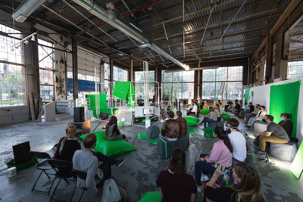 A seated group of people looking at a lecture on a monitor with bright green fabric and pillows spread throughout the space