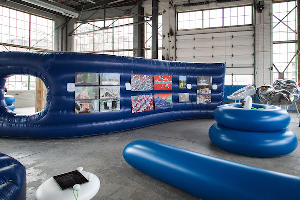 An installation of bright blue inflatable objects. One is a wavy wall with art hung on it.
