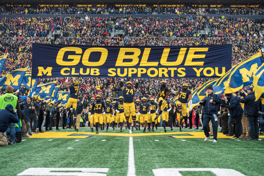 The Michigan Wolverines entering Michigan Stadium under a banner that says Go Blue, M Club Supports You