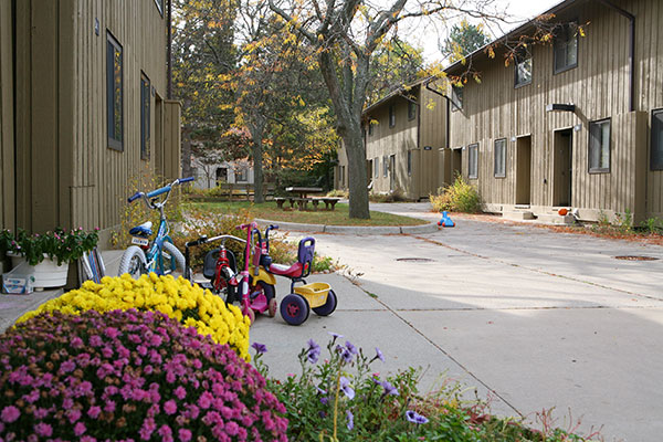 A walkway between wooden apartment buildings with colorful mums and kids' bikes