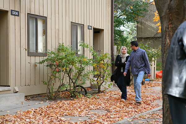 A couple walking in front of an apartment building with autumn leaves on the ground. The woman is wearing a hijab.