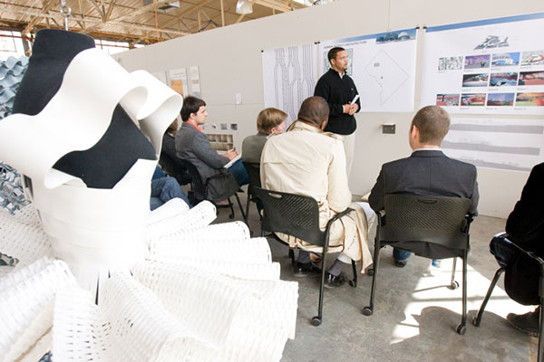 An architecture student defending a thesis in front of a group of people. There are thesis posters on the wall and architectural models.